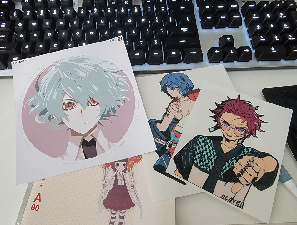 Three vinyl anime stickers on a desk with a notebook with a sticker on the front.